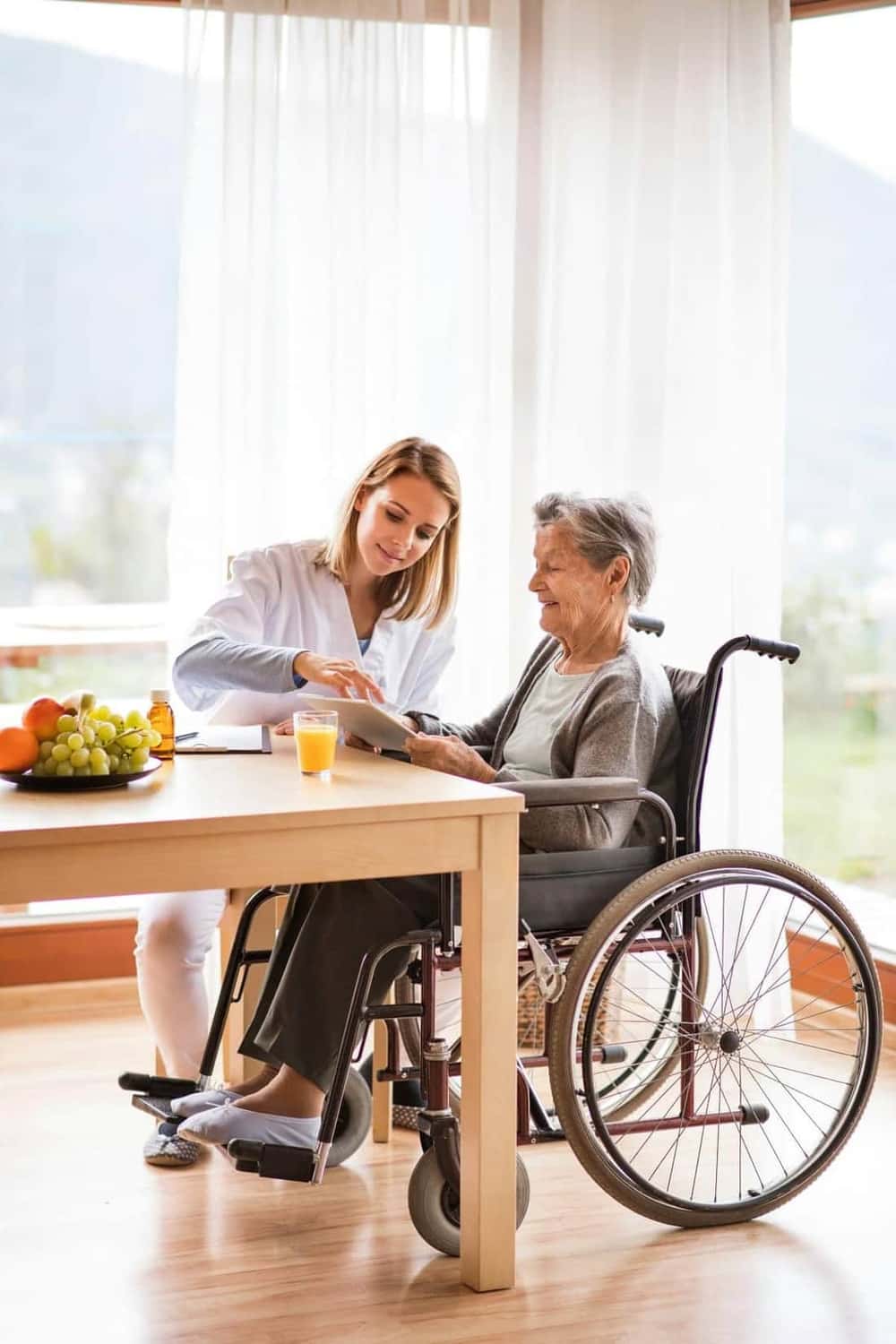 A young caregiver sitting beside an older woman in a wheelchair at a sunlit table — a calm scene representing thoughtful planning during accelerating care needs
