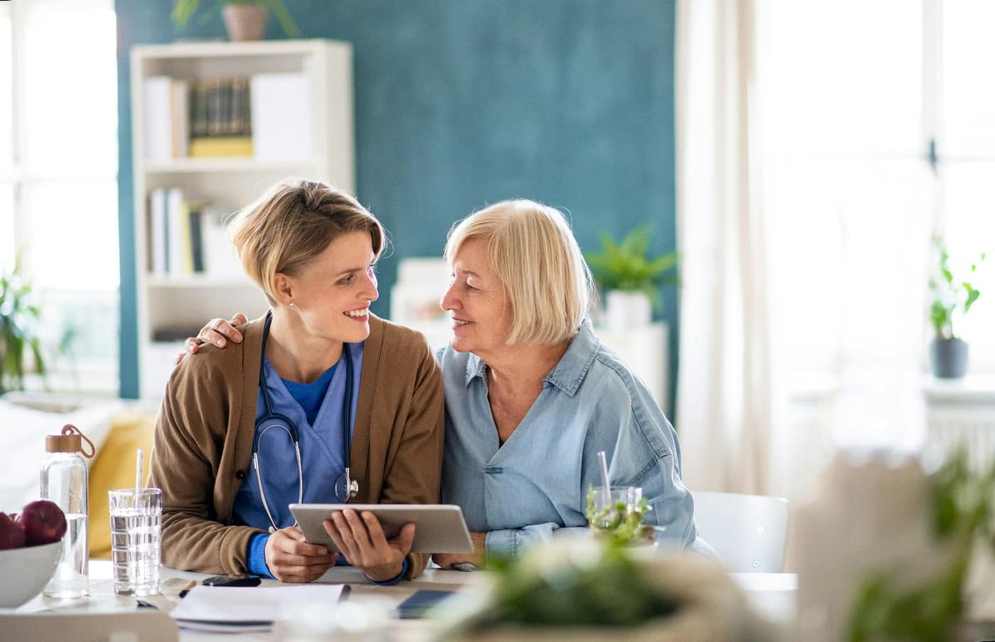 A nurse and an older woman sharing a warm moment at a sunlit table with a tablet — representing connection and thoughtful Alzheimer's care planning