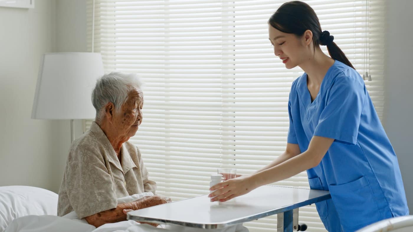 A professional caregiver in scrubs gently helping an older woman with medication at her bedside — a calm scene representing attentive full-time care