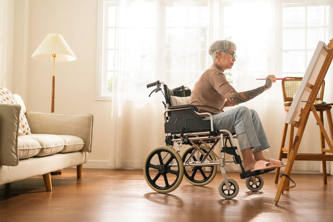Older woman in a wheelchair painting at an easel in a sunlit living room — a calm scene representing continued meaningful activity during cognitive change