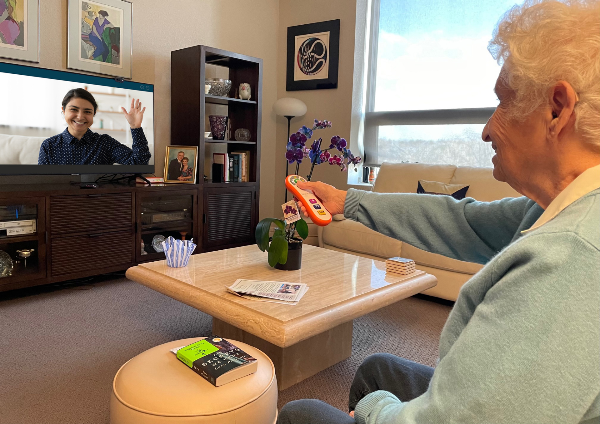 An older woman using the OdessaConnect orange universal remote to video-call her granddaughter on the TV