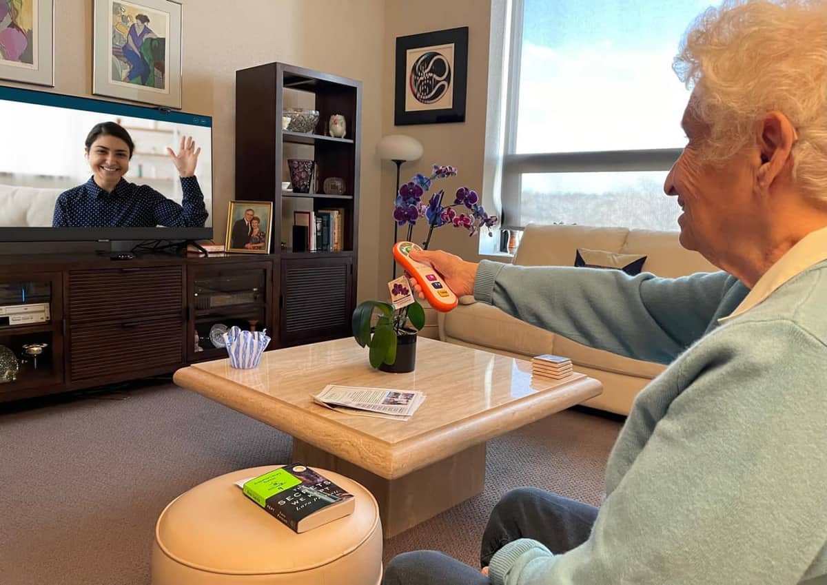 An older woman using the OdessaConnect orange universal remote to video-call her granddaughter on the TV