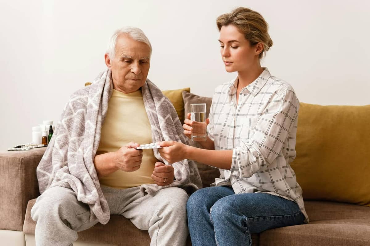 A daughter helps her father with medication on the couch — representing thoughtful at-home support for a loved one with Parkinson's